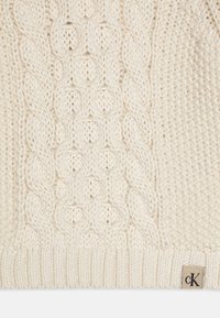 Pull en maille torsadée de couleur crème avec des motifs texturés et un ourlet côtelé. Étiquette de marque brodée visible en bas à droite.