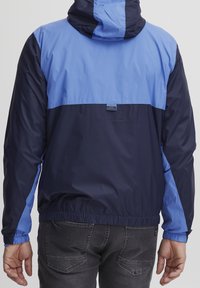 Veste coupe-vent avec panneaux bleu marine et bleu clair. Comprend une capuche, des poignets élastiques et un patch logo au dos. Matière légère.