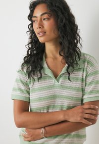 Sieviete ar cirtainiem melniem matiem, valkājot zaļu un baltu svītrainu īsroku adītu kreklu ar apkakli, krusto rokas un skatās pa kreisi.