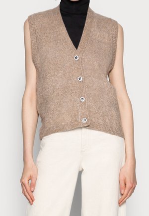 Gilet en maille beige sans manches avec quatre boutons floraux porté sur un col roulé noir et associé à un pantalon blanc cassé.