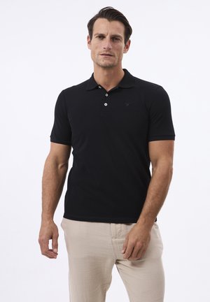 Camisa polo negra hecha de una tela suave, con un cierre de tres botones y un logo sutil. Combinada con pantalones de color beige claro.