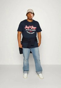 Navy cotton t-shirt with red and white logo, light blue denim jeans, and white sneakers. A dark sweater is tied around the waist, cotton cap worn.