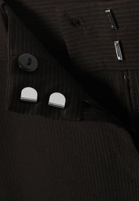 Tissu texturé brun foncé avec un bouton noir, deux clips métalliques sur le rabat et une fermeture éclair partiellement visible sur le côté.