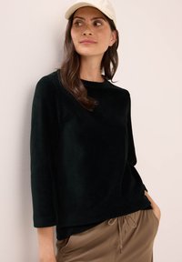 Vrouw in een crèmekleurige pet, zwart langwerpig ribtop en bruine broek met trekkoord, staand tegen een lichte muur, met de blik opzij.