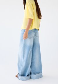 Jean denim à jambes larges de couleur bleu clair avec un ourlet brut et des bords retroussés, associé à un haut court jaune vif. Texture lisse.