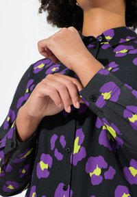 Sort skjorte med lilla og limegrøn blomsterdesign, med knaplukning og lange ærmer. Manchetter ved håndleddet med synlige knapdetaljer.
