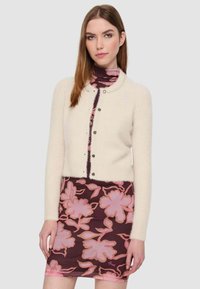 Cardigan color crema con scollo rotondo, chiusura a bottoni e tessuto texturizzato, indossato sopra un abito a fantasia floreale scuro.