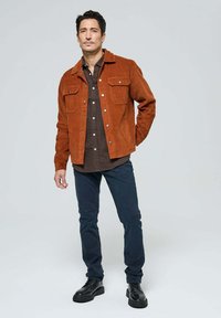 Homme debout avec une main dans la poche portant une veste en velours côtelé rouille, une chemise à carreaux marron, un pantalon bleu marine et des chaussures en cuir noir sur fond uni.