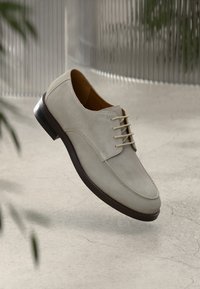 Ανοιχτό γκρι σουέτ παπούτσι με στρογγυλή μύτη, σχέδιο με κορδόνια και σκούρο καφέ σόλα. Λείο υλικό με ελάχιστες λεπτομέρειες.