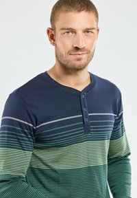 Homme aux cheveux courts portant une chemise henley à manches longues rayée bleu marine et verte avec trois boutons, regardant légèrement souriant.