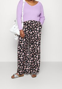Woman wearing lavender ribbed V-neck sweater, black wide-leg floral pants, white shoulder bag, and silver open-toe sandals standing against plain background.