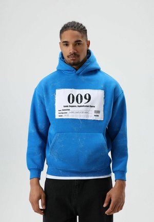 Sweat à capuche bleu avec poche avant et une grande étiquette blanche affichant le numéro "009" ainsi qu'un texte descriptif. Tissu doux avec légère usure.