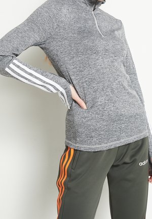 Person trägt ein graues Langarmshirt mit weißen Streifen an den Ärmeln und graue Adidas-Hosen mit orangenen Streifen und steht mit der Hand in der Hüfte.