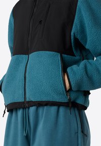 Giacca in pile teal con dettagli neri, chiusura con zip anteriore e orlo regolabile. Presenta una superficie testurizzata e tasche laterali.