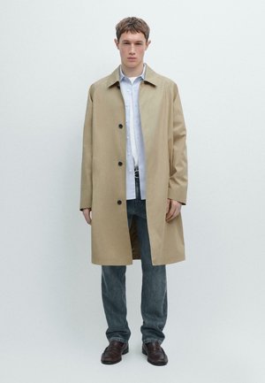 Mladý muž stojící, oblečený v béžovém kabátu po kolena, bílé košili na knoflíky, modrých džínách a tmavě hnědých botách, na jednotném bílém pozadí.