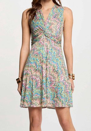 Bezrekawowa sukienka do kolan z żywym, wielokolorowym wzorem zygzaków oraz skręconym detalem na dekolcie w kształcie litery V przy talii, noszona z złotymi bransoletkami.