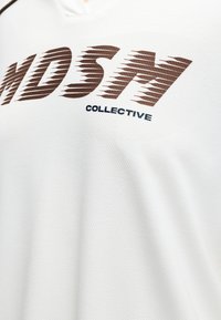 Tissu blanc texturé avec de grandes lettres marron "NDSH" au-dessus d'un texte noir plus petit "COLLECTIVE" sur une chemise.