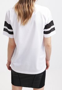 Person med långt hår som bär en vit kortärmad skjorta med svarta ränder på ärmarna och en svart kjol, vänd bort från kameran.