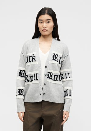 Ženska, oblečena v sivo kardigan z gumbi in črnim gotskim napisom na beli majici z V-izrezom ter rjavih hlačah, stoji pred enostavnim ozadjem.