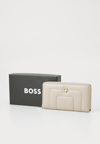 Portafoglio in pelle beige con chiusura a zip, caratterizzato da un design trapuntato e un dettaglio del logo in oro, posizionato accanto a una scatola nera con logo.