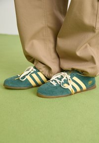 Tureckie zamszowe trampki w kolorze teal z trzema żółtymi paskami, białymi sznurowadłami i złotymi akcentami. Noszone z beżowymi spodniami na zielonej teksturowanej powierzchni.