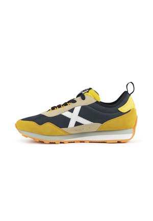 Zapatilla amarilla y negra con diseño blanco en forma de X en el lateral, cordones negros, detalles beige y suela de goma naranja, vista desde un lado.