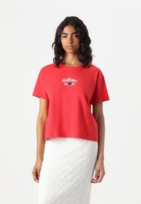 Red cotton t-shirt with a cropped design, featuring a white embroidered "Hollister 2002" logo on the front, paired with a textured white skirt.