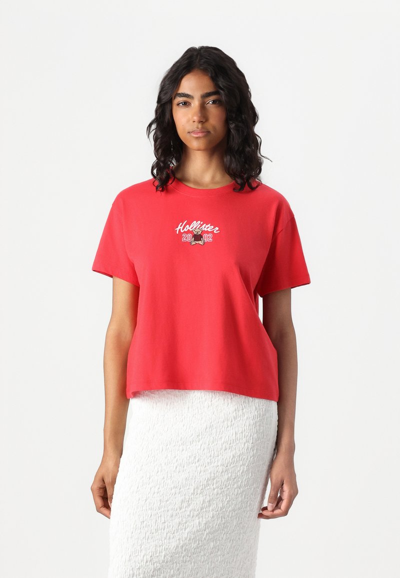 Röd bomullströja med kortad design, med en vit broderad "Hollister 2002" logotyp på framsidan, tillsammans med en texturerad vit kjol.