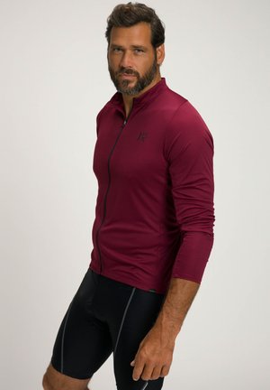 Jersey de ciclismo de manga larga en burdeos, hecho de tejido texturizado con cremallera frontal y logo negro, combinado con pantalones cortos de ciclismo negros.