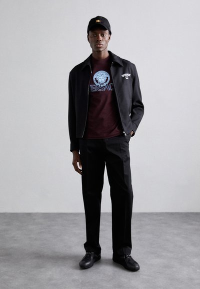 Sort zip-up jakke over en bordeaux T-shirt med et blåt Versace grafik, parret med sorte bukser og sorte sko; kasket med logo.