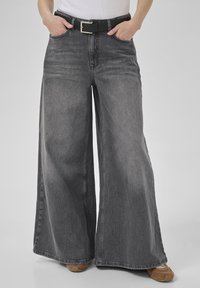 Person wearing high-waisted, wide-leg faded black jeans with a black belt, white top, and brown sneakers, hands in pockets.