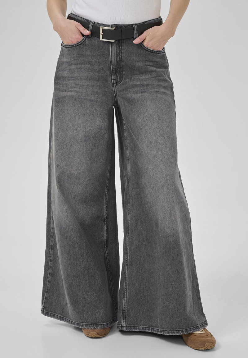 Person wearing high-waisted, wide-leg faded black jeans with a black belt, white top, and brown sneakers, hands in pockets.