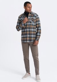 Ruitjes flanellen shirt in blauw, geel en grijs tinten, met knoopsluiting aan de voorkant, twee borstzakken en een gebogen zoom. Gecombineerd met beige broek en sneakers.