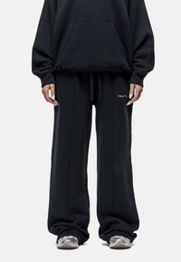 Zwarte sweatshirt met een voorzak, gecombineerd met losse zwarte joggingbroeken met een trekkoord en zijnaaddetails, gedragen met grijze sneakers.