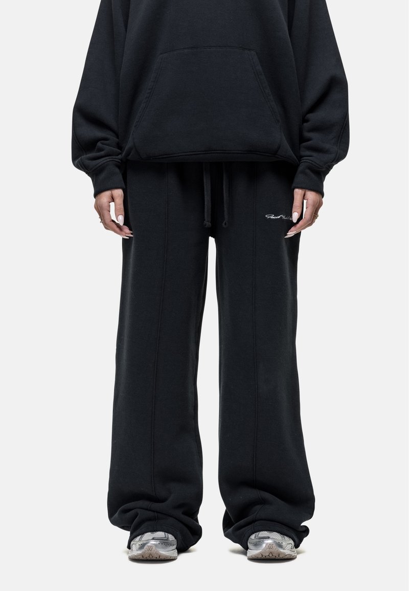 Zwarte sweatshirt met een voorzak, gecombineerd met losse zwarte joggingbroeken met een trekkoord en zijnaaddetails, gedragen met grijze sneakers.