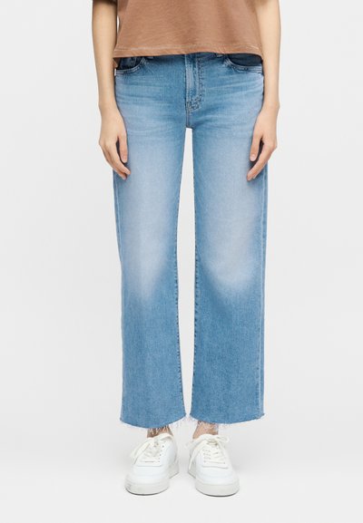 Άτομο που φοράει ανοιχτό μπλε τζιν με ίσια γραμμή και ξεφτισμένα τελειώματα, λευκά αθλητικά παπούτσια και καφέ μπλούζα, στέκεται μπροστά σε απλό φόντο.