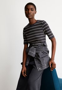 Zwart-wit gestreepte top met korte mouwen, gecombineerd met een geknoopte grijze spijkerjack, gedragen over donkere, comfortabele broek, tegen een effen achtergrond.