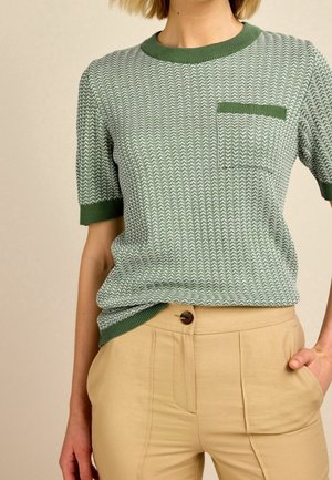 Femme portant un pull à manches courtes à motif vert et blanc avec bordure verte et pantalon beige taille haute, main dans la poche.