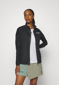 Schwarze leichte Jacke mit hohem Kragen, Reißverschluss vorne und Terrex-Logo, kombiniert mit einem weißen T-Shirt und grünen Shorts. Glatte Textur.