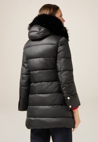 Abrigo acolchado negro con cuello de piel sintética, con acolchado horizontal y detalles de botones a presión en las mangas. Acabado suave y brillante.