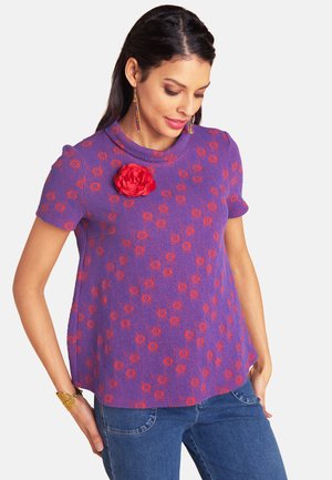 Frau trägt ein lila strukturiertes Oberteil mit orangefarbenem Blumenmuster und einer roten Stoffblumen-Brosche, kombiniert mit blauer Jeans und bunten Ohrringen.