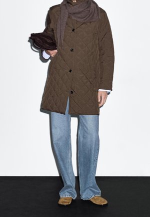 Henkilö, jolla on päällään ruskea tikattu takki, vaaleansiniset farkut ja vaaleanruskeat kengät, ja joka pitää käsissään tummanruskeaa hattua sekä käyttää ruskeaa huivia.