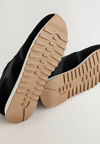 Schwarze Sneaker mit beigen Gummisohlen und tiefem Profil, aus einem schrägen Blickwinkel gezeigt, der die strukturierte Außensohle hervorhebt.