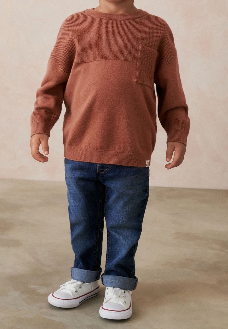 Enfant portant un pull couleur rouille avec une poche devant, un jean bleu retroussé et des baskets blanches debout sur un sol beige.