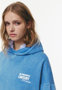 Felpa blu in pile con un design over-size, caratterizzata da un ampio cappuccio e il testo del logo bianco "MID CENTURY Art Studio". Colletto a righe della camicia visibile sotto.