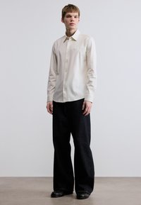 Ανοιχτό κρεμ πουκάμισο με κουμπιά και γιακά, σε συνδυασμό με άνετο σκούρο μπλε τζιν και μαύρα παπούτσια, που στέκεται σε ουδέτερο φόντο.