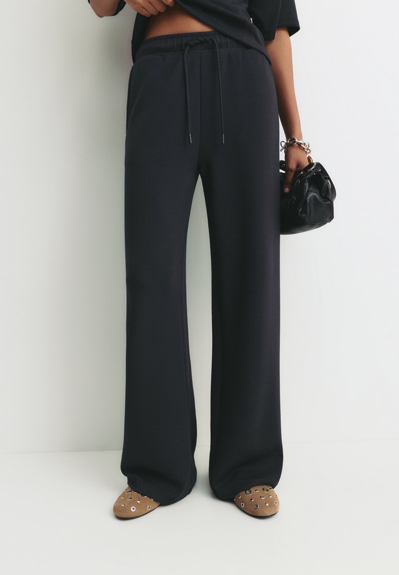 Personne portant un pantalon noir à jambes larges, tenant un petit sac à main noir, avec des chaussures marron cloutées visibles sous le pantalon.