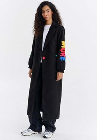 Kobieta z długimi kręconymi włosami nosi czarny długi płaszcz z kolorowym napisem "MORE" na rękawie, białą koszulę, czarne spodnie i białe tenisówki.