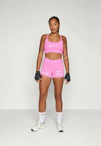 Rózsaszín sportos crop top és rövidnadrág, fehér Nike logókkal. Az öltözék testhezálló, sima tapintású. Fekete kesztyűk és fehér zoknik egészítik ki a megjelenést.