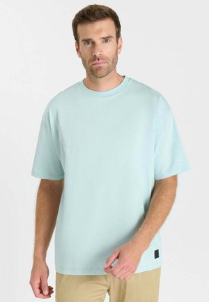 Mann mit kurzen braunen Haaren, der ein hellblaues übergroßes T-Shirt und beige Hosen trägt, vor einem weißen Hintergrund stehend.